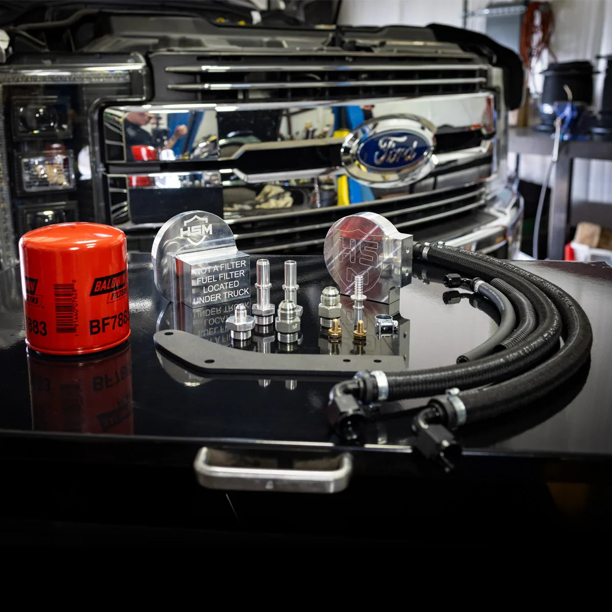 H&S Motorsports 2011–2016 fuel filter relocation kit displayed on a workbench in front of a Ford Super Duty grille, showing all components including hoses, billet blocks, fittings, and Baldwin filter.