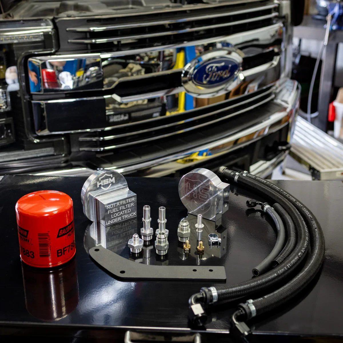 Full H&S Motorsports fuel filter relocation kit for 2017–2023 Ford 6.7L Power Stroke displayed in front of Ford Super Duty grille, including billet adapters, fittings, hoses, and red Baldwin filter.