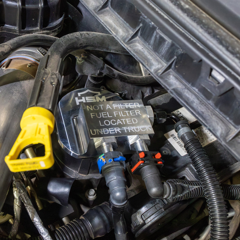 
                  
                    Installed view of HSM fuel filter bypass kit on 2011–2016 Ford 6.7L Power Stroke engine, showing billet block and connected quick-disconnect fuel lines.
                  
                
